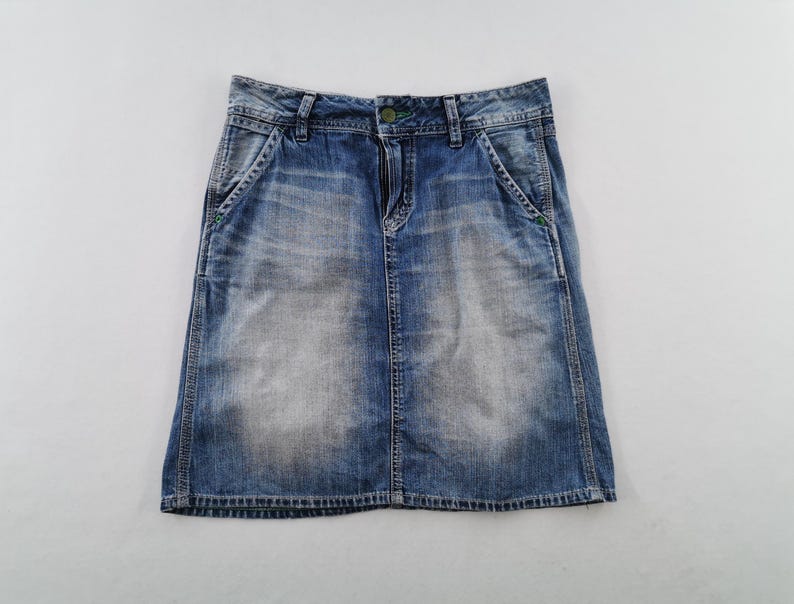 Pu&ograve; includere: Una gonna di jeans blu con un lavaggio sbiadito. La gonna ha un classico design a cinque tasche, una chiusura con bottone e cerniera e cuciture verdi sulle tasche. La gonna &egrave; lunga circa 46 cm.