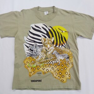 Puede incluir: Camiseta color beige con un leopardo, cebras y un pájaro sobre un fondo de rayas de cebra y un sol amarillo. Incluye la palabra "SINGAPORE" y la firma del artista. La camiseta está hecha de un material suave y cómodo.