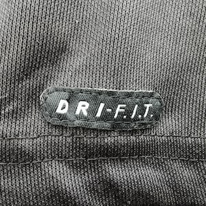 Pu&ograve; includere: Primo piano di un tessuto grigio scuro con una cucitura. Una toppa rettangolare con la scritta "DRI-FIT" in lettere bianche &egrave; cucita sul tessuto. Il tessuto ha un aspetto strutturato.