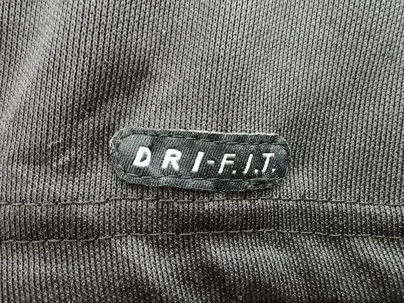 Pu&ograve; includere: Primo piano di un tessuto grigio scuro con una cucitura. Una toppa rettangolare con la scritta "DRI-FIT" in lettere bianche &egrave; cucita sul tessuto. Il tessuto ha un aspetto strutturato.