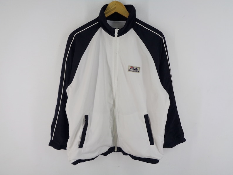 May include: A white and navy blue track jacket with a full zipper closure. The jacket features a high collar, side pockets, and the "FILA" logo on the chest. White piping accents the navy blue sleeves.