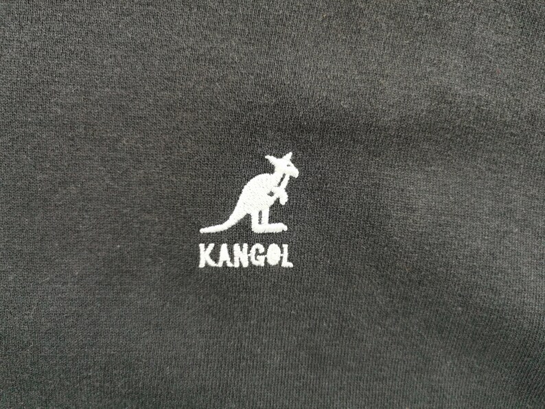 May include: Black fabric with a white embroidered kangaroo logo and the brand name "KANGOL" below it. The logo is a stylized kangaroo holding a cane. The fabric appears to be a t-shirt or other apparel item.