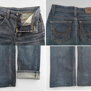 K&ouml;nnte beinhalten: Dunkelblaue Jeans aus Denim mit Rei&szlig;verschluss und Knopf, zwei Ges&auml;&szlig;taschen mit goldfarbenen N&auml;hten und einem braunen Lederaufn&auml;her am Bund mit der Aufschrift Edwin. Die Beinhosen sind gerade geschnitten, ein Bein ist umgeschlagen.