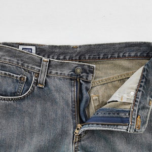 May include: Close-up of a pair of faded blue denim jeans with a partially unzipped fly. The jeans have a classic five-pocket design, a button closure, and belt loops. The brand name "EDWIN" is visible on a small tag.