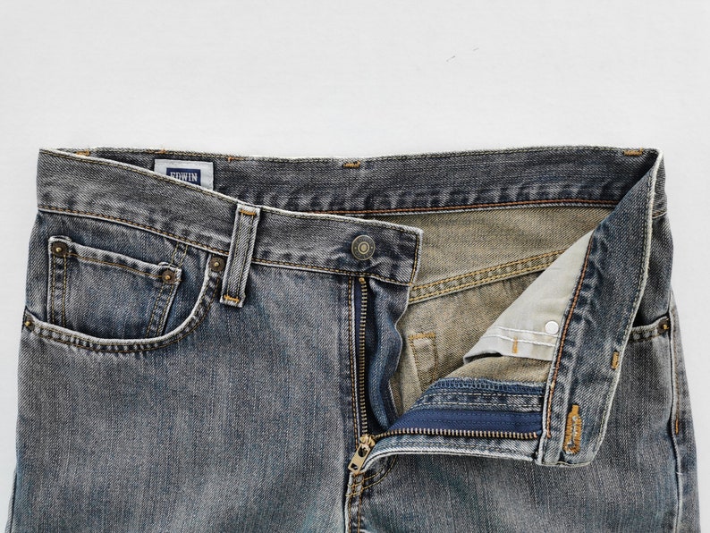 May include: Close-up of a pair of faded blue denim jeans with a partially unzipped fly. The jeans have a classic five-pocket design, a button closure, and belt loops. The brand name "EDWIN" is visible on a small tag.