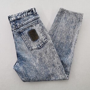 May include: Acid wash denim jeans with a high waist and a straight leg. The jeans have a classic five-pocket design, a button and zipper closure, and a brown leather patch on the back pocket. The denim has a faded, distressed look.