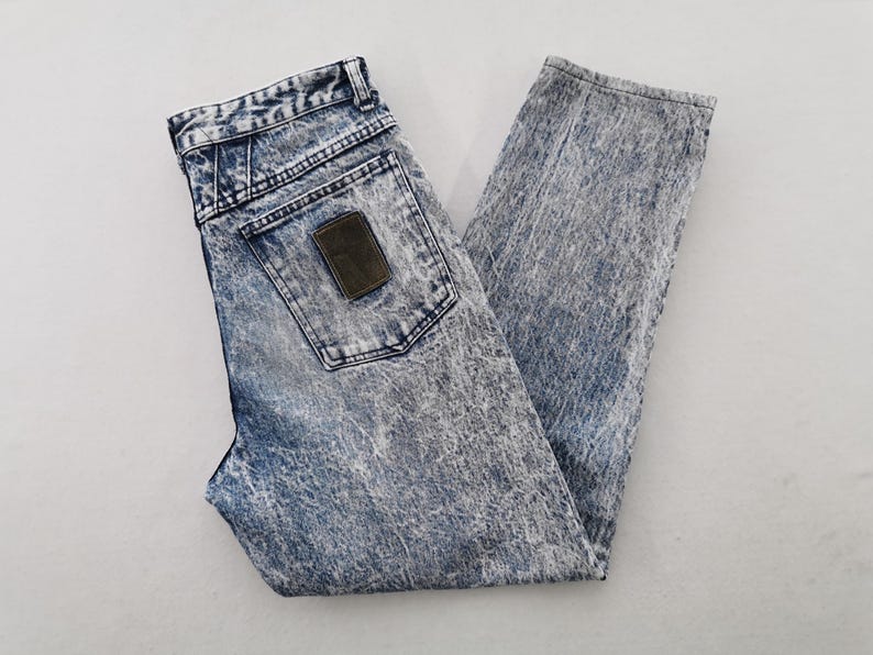 May include: Acid wash denim jeans with a high waist and a straight leg. The jeans have a classic five-pocket design, a button and zipper closure, and a brown leather patch on the back pocket. The denim has a faded, distressed look.