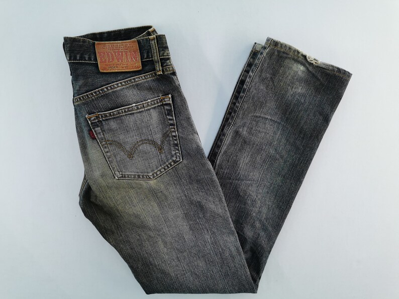 May include: Faded black denim jeans with a classic five-pocket design. Features a leather patch with the brand name "EDWIN" and a red logo on the back pocket. The jeans are folded and laid flat.