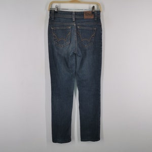K&ouml;nnte beinhalten: Eine dunkelblaue Jeans aus Denim mit geradem Bein, zwei Ges&auml;&szlig;taschen mit dekorativen goldfarbenen N&auml;hten, einem braunen Leder-Markenaufn&auml;her am Bund und einem klassischen F&uuml;nf-Taschen-Design mit Metallnieten und einem Knopfverschluss.