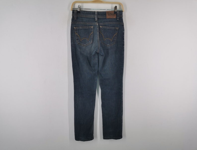 K&ouml;nnte beinhalten: Eine dunkelblaue Jeans aus Denim mit geradem Bein, zwei Ges&auml;&szlig;taschen mit dekorativen goldfarbenen N&auml;hten, einem braunen Leder-Markenaufn&auml;her am Bund und einem klassischen F&uuml;nf-Taschen-Design mit Metallnieten und einem Knopfverschluss.