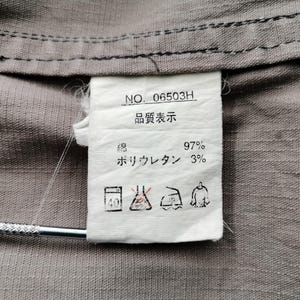 May include: Close-up of a gray fabric with a white tag attached. The tag has text in Japanese and English, including "NO. 06503H" and fabric composition details. The fabric appears to be a blend of cotton and polyurethane.