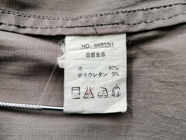 May include: Close-up of a gray fabric with a white tag attached. The tag has text in Japanese and English, including "NO. 06503H" and fabric composition details. The fabric appears to be a blend of cotton and polyurethane.
