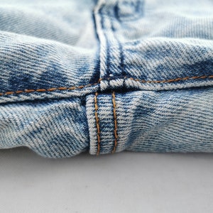 May include: Close-up of light blue denim jeans. The image highlights the seam details, with visible orange stitching and the texture of the fabric. The jeans appear to be well-worn.