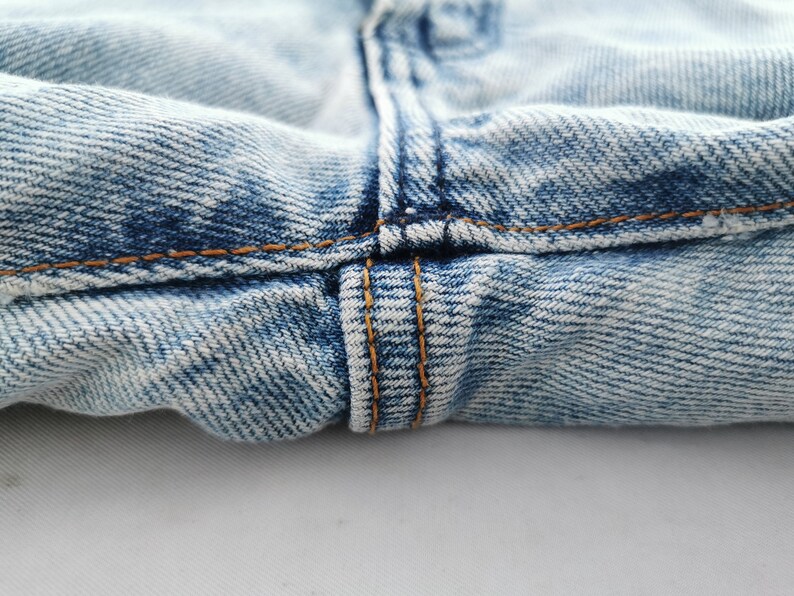 May include: Close-up of light blue denim jeans. The image highlights the seam details, with visible orange stitching and the texture of the fabric. The jeans appear to be well-worn.