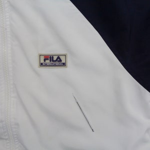 May include: White and navy blue athletic jacket with a full zipper. The jacket features the "FILA INTERNATIONAL" logo patch on the chest. The jacket has a color block design.