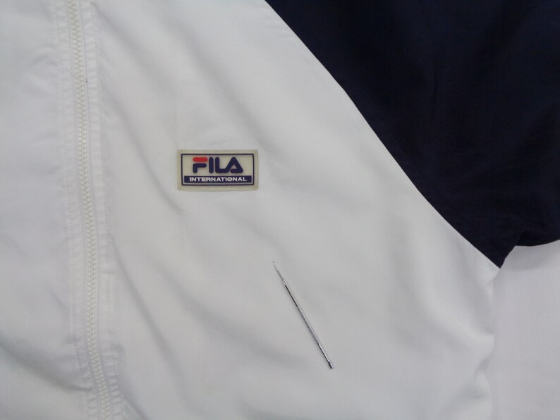 May include: White and navy blue athletic jacket with a full zipper. The jacket features the "FILA INTERNATIONAL" logo patch on the chest. The jacket has a color block design.