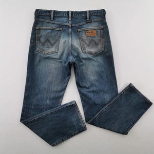 May include: Blue denim jeans with a classic five-pocket design. The jeans feature a faded wash and the Wrangler logo patch on the back. The jeans are laid flat on a white surface.