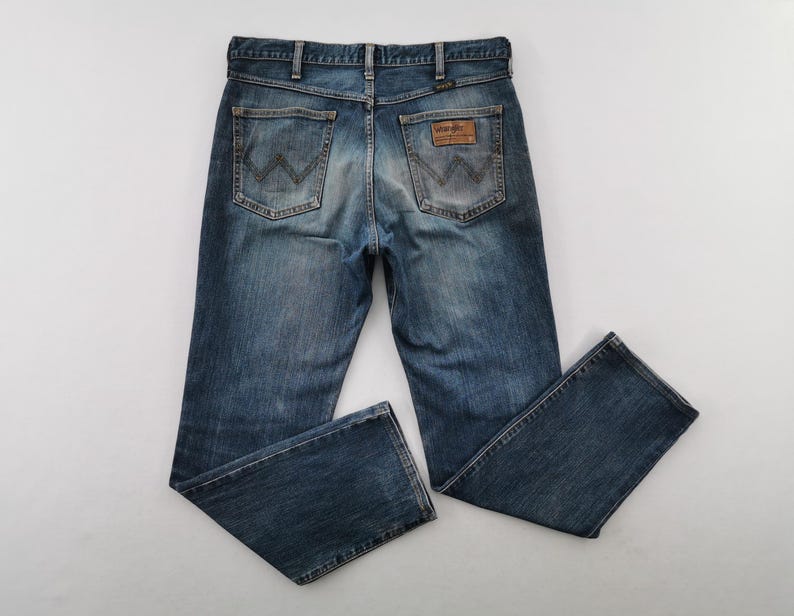 May include: Blue denim jeans with a classic five-pocket design. The jeans feature a faded wash and the Wrangler logo patch on the back. The jeans are laid flat on a white surface.