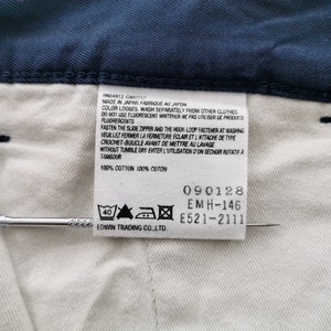 May include: Close-up of a navy blue garment with a white interior and a tag. The tag includes text in English and French, along with care symbols and the text "100% COTTON". A silver pin is placed near the tag.