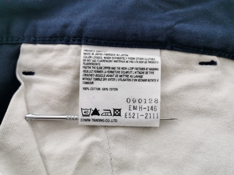 May include: Close-up of a navy blue garment with a white interior and a tag. The tag includes text in English and French, along with care symbols and the text "100% COTTON". A silver pin is placed near the tag.