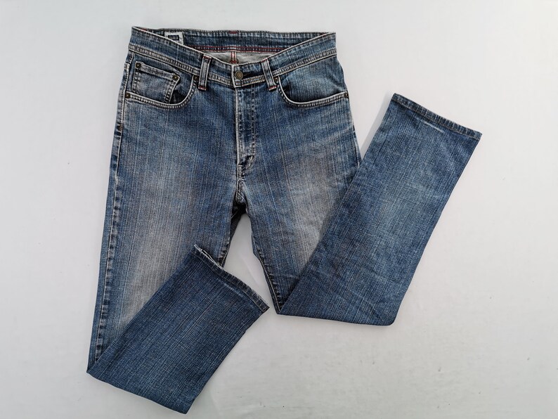 May include: Blue denim jeans with a classic five-pocket design. The jeans have a button and zip closure, belt loops, and a faded wash. The legs are straight cut. The jeans are laid flat on a white surface.