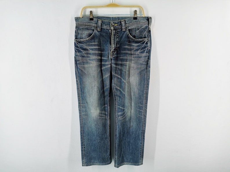 May include: Blue denim jeans hanging on a gold hanger. The jeans have a classic five-pocket design and a straight leg cut. The denim has a faded, worn look with lighter areas and subtle distressing.