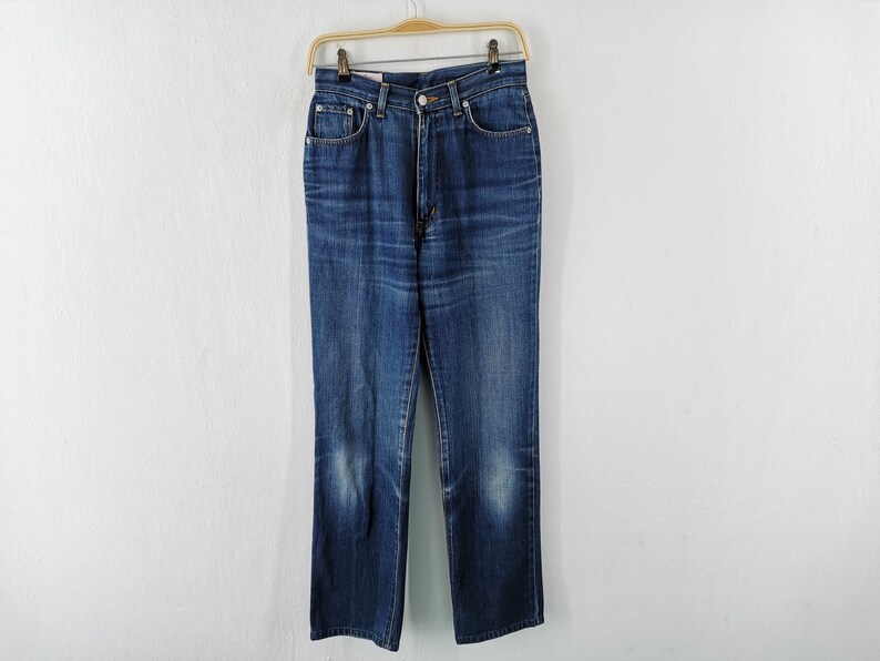 May include: A pair of blue denim jeans hanging on a wooden hanger. The jeans have a classic five-pocket design, a button and zipper closure, and a straight leg. The denim has a faded appearance.
