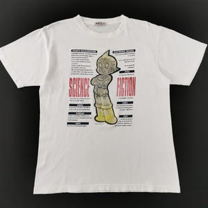 May include: A white short-sleeve t-shirt featuring a central graphic of the character Astro Boy in yellow and black. The design includes the text SCIENCE FICTION in red and various technical descriptions in black text surrounding the character.