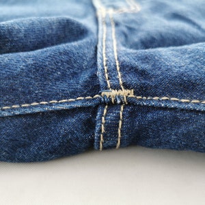 May include: Close-up of blue denim jeans, showcasing the seam details. The stitching is a contrasting light beige color, highlighting the construction of the garment. The fabric has a textured appearance.