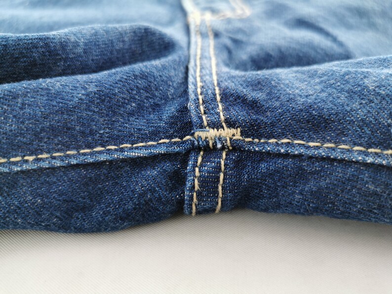 May include: Close-up of blue denim jeans, showcasing the seam details. The stitching is a contrasting light beige color, highlighting the construction of the garment. The fabric has a textured appearance.