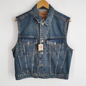 May include: A sleeveless denim vest in a medium blue wash, with a button-up front, two chest pockets, and a collar. The vest has a "BIG JOHN" label and a price tag attached. The vest is hanging on a wooden hanger.