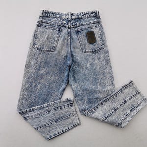 May include: Acid wash denim jeans with a high waist and a classic five-pocket design. The jeans have a faded blue and white pattern, with a brown leather patch on the back. The jeans are folded and laid flat.