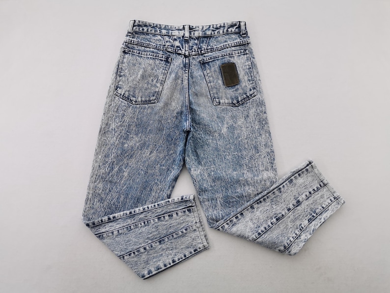 May include: Acid wash denim jeans with a high waist and a classic five-pocket design. The jeans have a faded blue and white pattern, with a brown leather patch on the back. The jeans are folded and laid flat.
