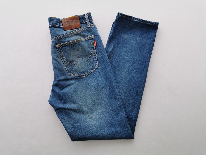 Puede incluir: Vaqueros de mezclilla azules con un dise&ntilde;o cl&aacute;sico de cinco bolsillos. Cuenta con un parche de cuero marr&oacute;n en la cintura trasera y una peque&ntilde;a etiqueta naranja. Los vaqueros est&aacute;n planos, mostrando el lavado descolorido y los detalles de costura.