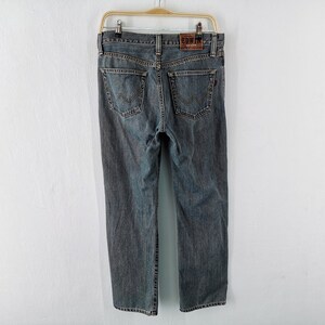 May include: A pair of gray denim jeans hanging on a wooden hanger. The jeans have a classic five-pocket design with a leather patch on the back that reads "EDWIN". The jeans are a medium wash and appear to be in good condition.