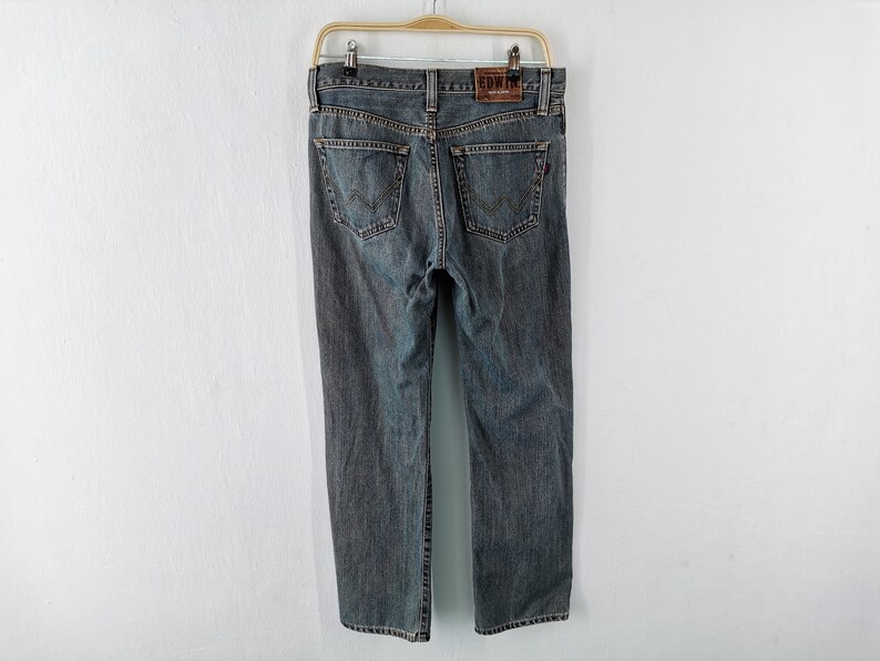 May include: A pair of gray denim jeans hanging on a wooden hanger. The jeans have a classic five-pocket design with a leather patch on the back that reads "EDWIN". The jeans are a medium wash and appear to be in good condition.