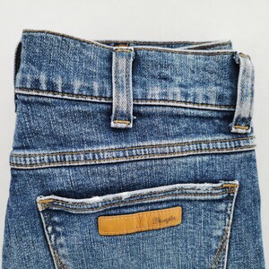 May include: Close-up of blue denim jeans with a light brown leather patch on the back pocket. The patch has the brand name "Wrangler" embossed on it. The jeans have a classic five-pocket design and a button and zipper closure.