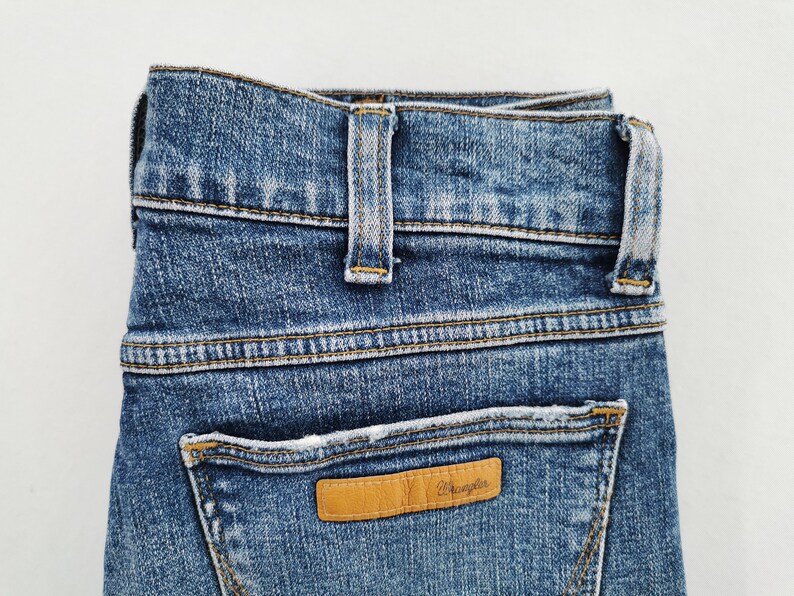 May include: Close-up of blue denim jeans with a light brown leather patch on the back pocket. The patch has the brand name "Wrangler" embossed on it. The jeans have a classic five-pocket design and a button and zipper closure.