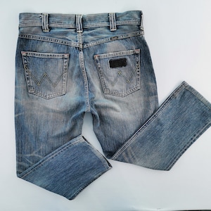 May include: Blue denim jeans with a classic five-pocket design. The jeans feature a faded wash, a leather patch, and the Wrangler logo on the back pockets. The jeans are folded and laid flat on a white surface.