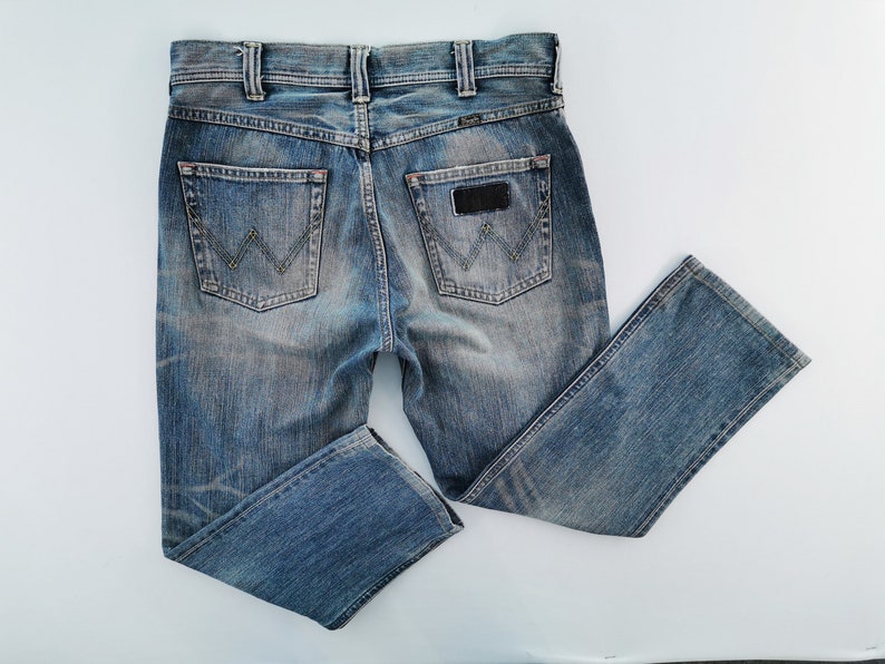May include: Blue denim jeans with a classic five-pocket design. The jeans feature a faded wash, a leather patch, and the Wrangler logo on the back pockets. The jeans are folded and laid flat on a white surface.