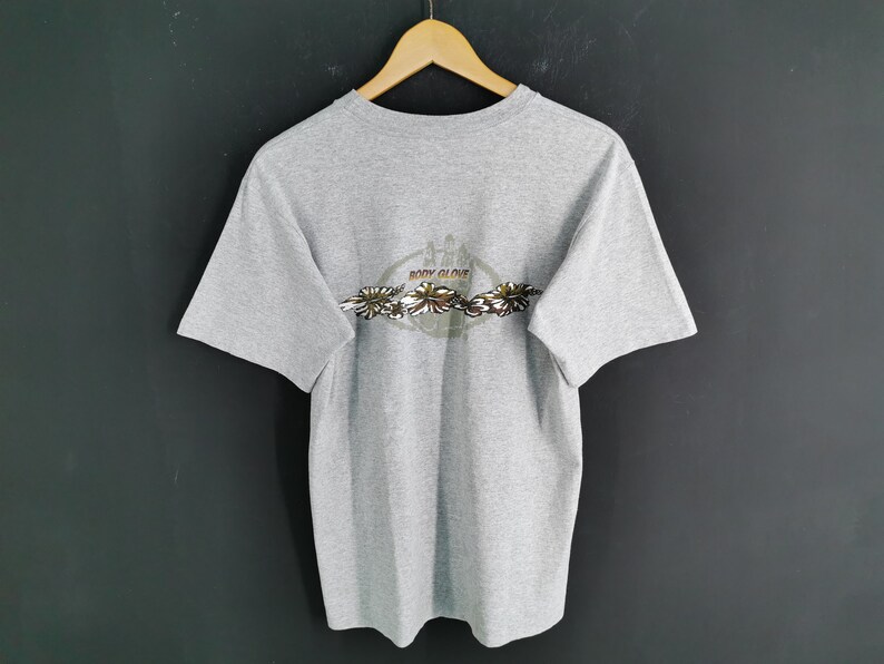 May include: A heather gray t-shirt with a crew neck and short sleeves. The back of the shirt features a circular design with the words "BODY GLOVE" and a floral pattern in shades of brown, green, and white.