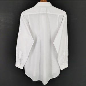 May include: A white long-sleeve shirt hanging on a wooden hanger. The shirt is made of a crisp, white fabric with a button-down collar and cuffs. The back of the shirt is visible, showing the tailored design and seams. The background is a dark gray.