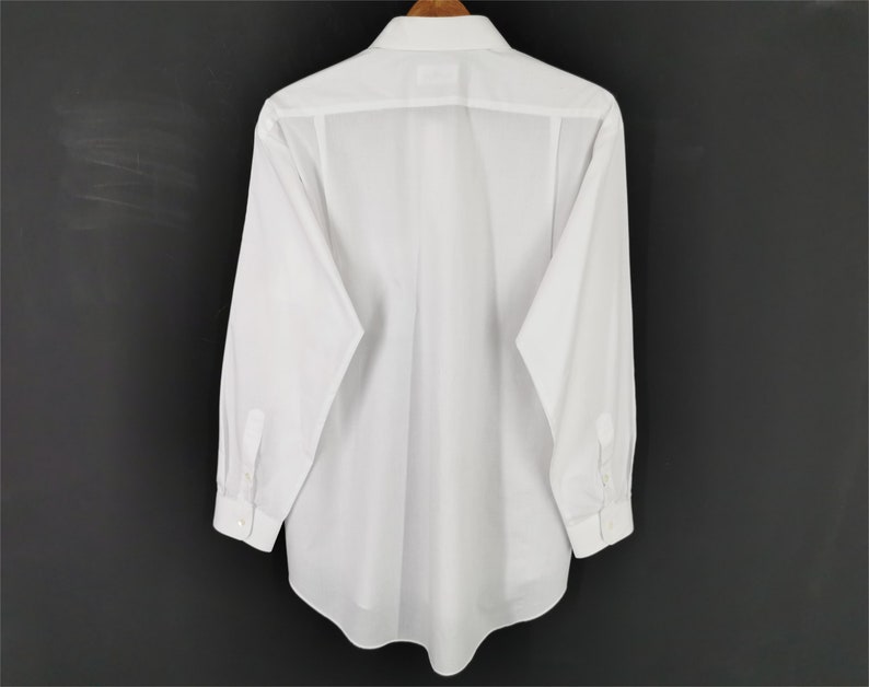May include: A white long-sleeve shirt hanging on a wooden hanger. The shirt is made of a crisp, white fabric with a button-down collar and cuffs. The back of the shirt is visible, showing the tailored design and seams. The background is a dark gray.