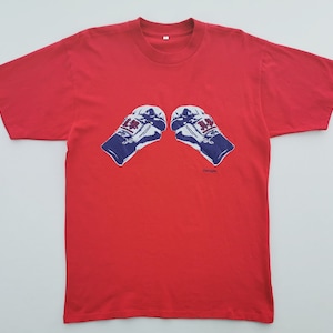 May include: A bright red short-sleeved t-shirt featuring a graphic print of two white and blue boxing gloves positioned in the center. The gloves have red accents and the text "Cologne" is printed in small blue letters below the right glove.