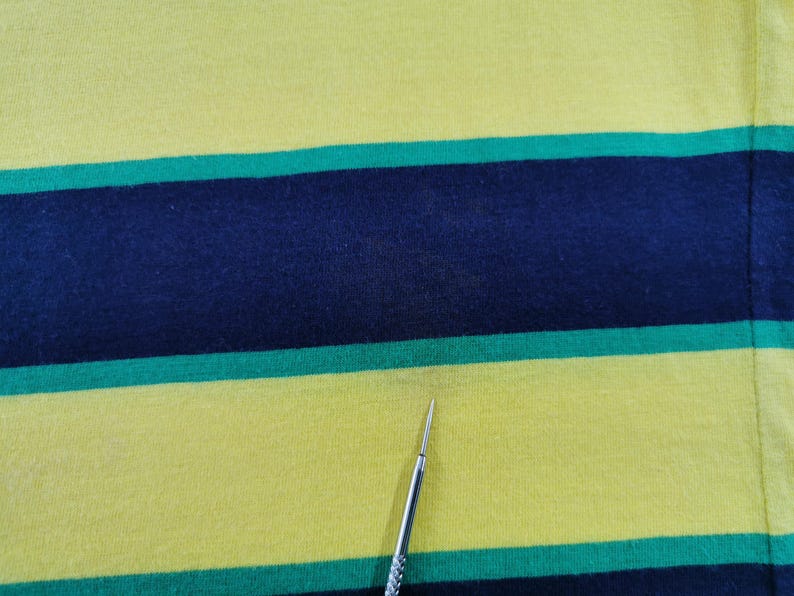 Op de afbeelding: Close-up van een gele stof met horizontale strepen in marineblauw en groen. De stof lijkt een gebreid materiaal te zijn, mogelijk katoen of een mix. De strepen zijn gelijkmatig verdeeld en cre&euml;ren een gedurfd, grafisch patroon.
