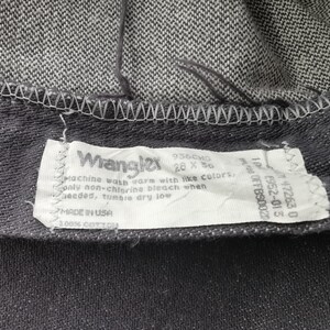 May include: Close-up of a pair of dark gray Wrangler jeans. The label reads "Wrangler" with size 28 x 36. The label also includes washing instructions and "MADE IN USA" text. The jeans are made of 100% cotton.