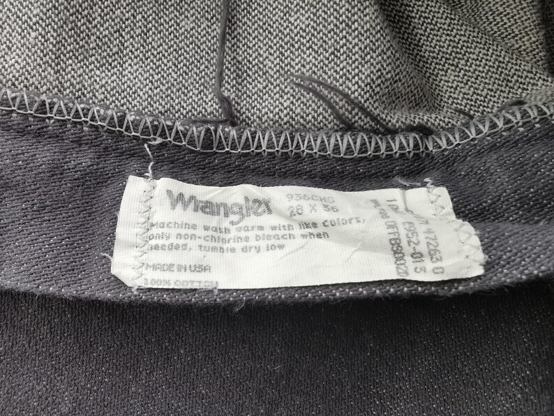 May include: Close-up of a pair of dark gray Wrangler jeans. The label reads "Wrangler" with size 28 x 36. The label also includes washing instructions and "MADE IN USA" text. The jeans are made of 100% cotton.