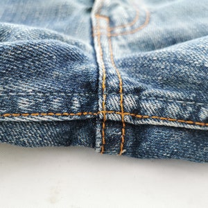 May include: Close-up of denim jeans, showcasing the detailed stitching and fabric texture. The image highlights the seams and the orange thread used in the construction of the garment. The denim is a classic blue wash.