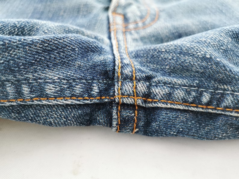 May include: Close-up of denim jeans, showcasing the detailed stitching and fabric texture. The image highlights the seams and the orange thread used in the construction of the garment. The denim is a classic blue wash.