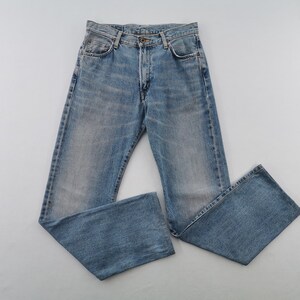 May include: Light blue denim jeans with a classic five-pocket design. The jeans have a button fly and a faded wash, giving them a vintage look. The jeans are laid flat on a light gray surface.